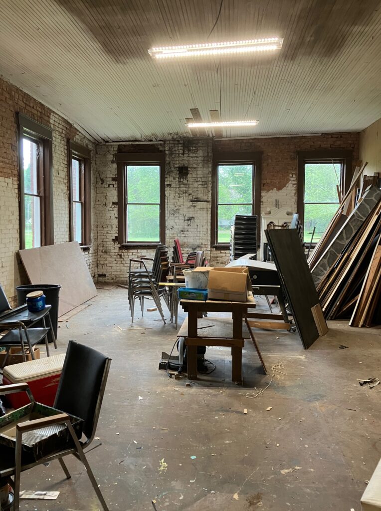 Workroom of the Crawford School shown with exposed brick walls and unpainted ceiling. Stacked chairs and scrap wood are scattered around.